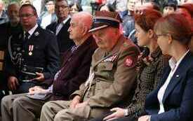 Uczestnicy uroczystości. Pośrodku siedzi kombatant w zielonym żołnierskim mundurze.