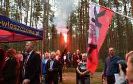 Młody człowiek  stoi z odpaloną czerwoną racą wok&oacute;ł inni uczestnicy uroczystości.
