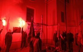 Przy symbolicznym grobie ks. Popiełuszki stoi poczet sztandarowy. Kilka os&oacute;b pali race.