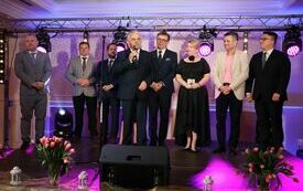 Organizatorzy i zaproszeni goście stoją na scenie i po kolei składają obecnym na sali paniom życzenia z okazji Dnia Kobiet