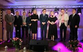 Organizatorzy i zaproszeni goście stoją na scenie i po kolei składają obecnym na sali paniom życzenia z okazji Dnia Kobiet