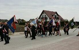 Poczty sztandarowe i uczestnicy uroczystości maszerują na cmentarz parafialny