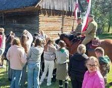Dzieci ze szkoły w Olesznie stoją przy dw&oacute;ch koniach. Jeźdźcy w strojach historycznych opowiadają dzieciom wojenne historie.