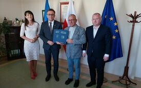 Starosta przekazuje księdzu jeden egzemplarz podpisanej umowy. Obok stoją Wicestarosa i Skarbnik Powiatu.