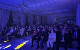 Publiczność siedzi na krzesłach przed sceną i słucha występu zespołu