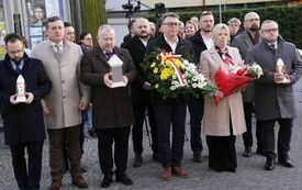 Na placu kościelnym stoją delegacje z wiązankami kwiat&oacute;w i zniczami