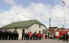 Wszyscy strażacy stoją na baczność, w tle widać w&oacute;z strażacki